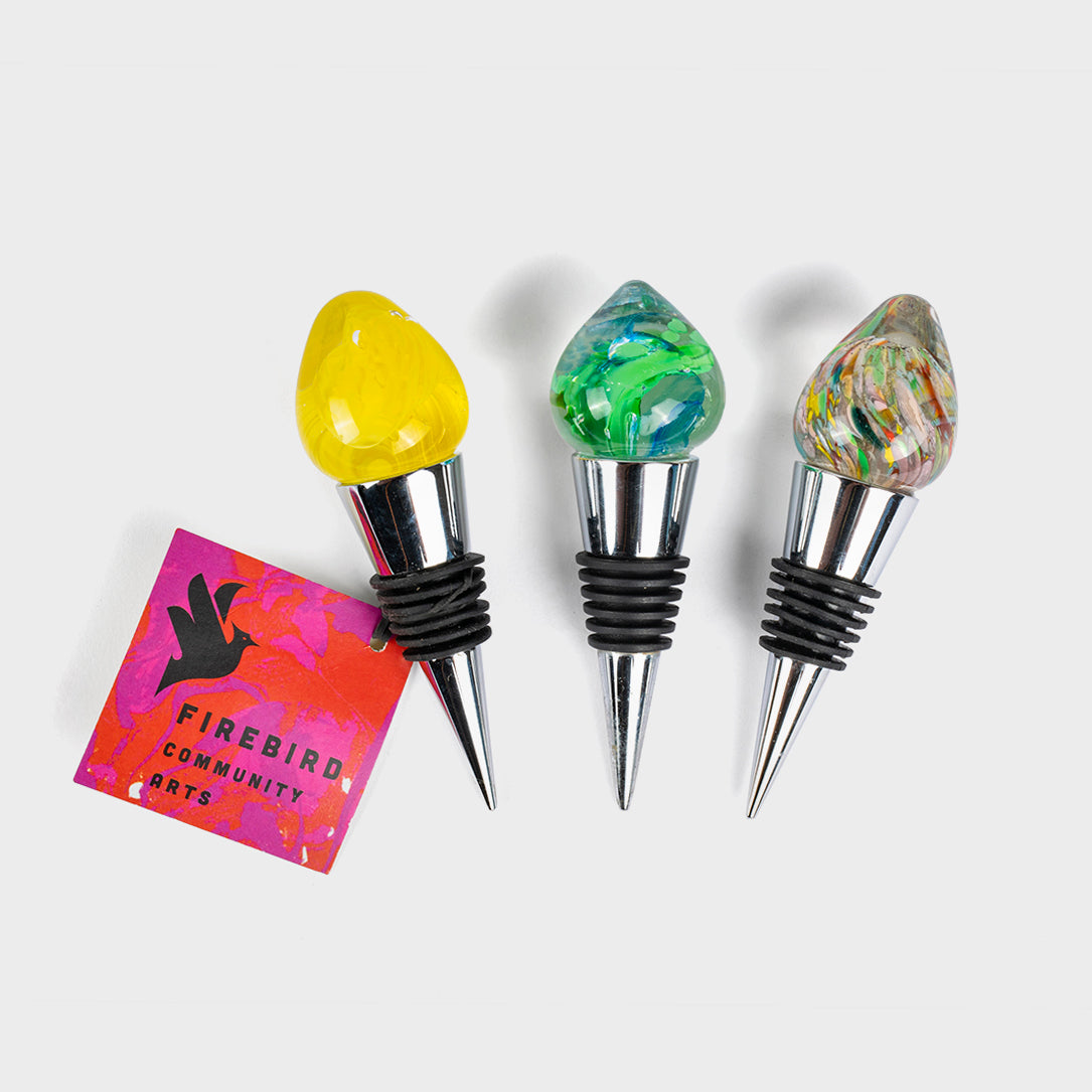 Three colorful glass wine stoppers with metal bases and rubber grips, handcrafted by Firebird Community Arts