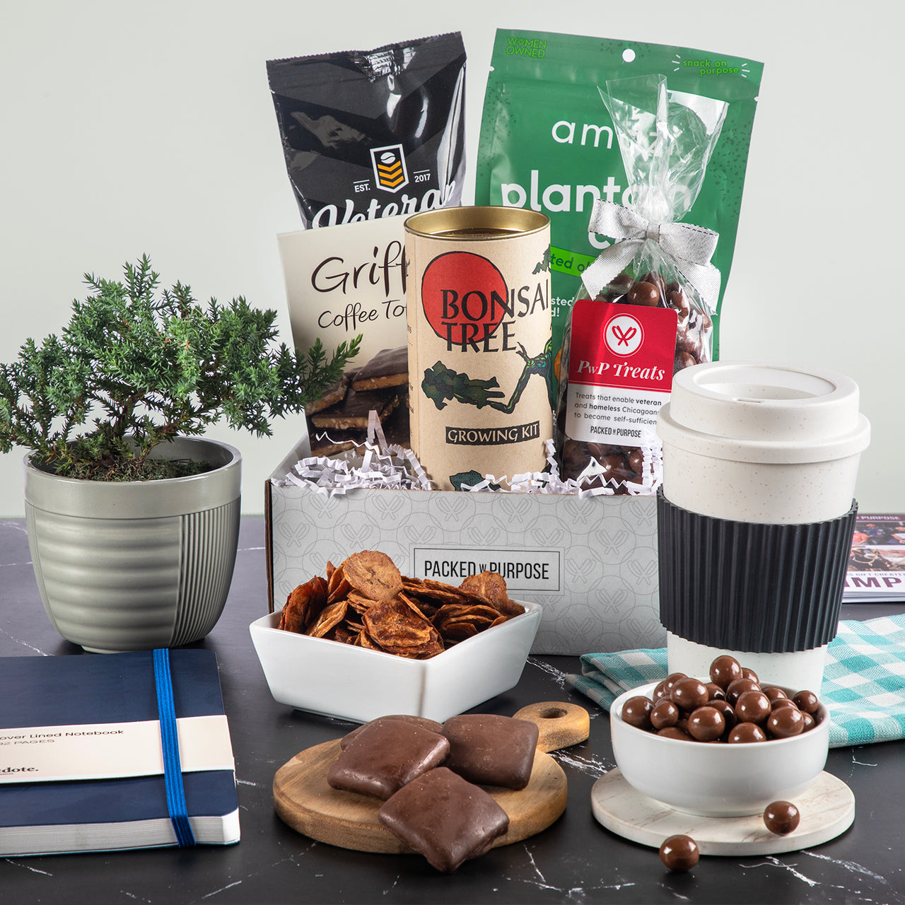 Open gift basket labeled ‘Positive Vibes Deluxe’ with terrazzo mug, organic tea, chocolate meltaways, jam, toffee, a journal and bonsai grow kit.
